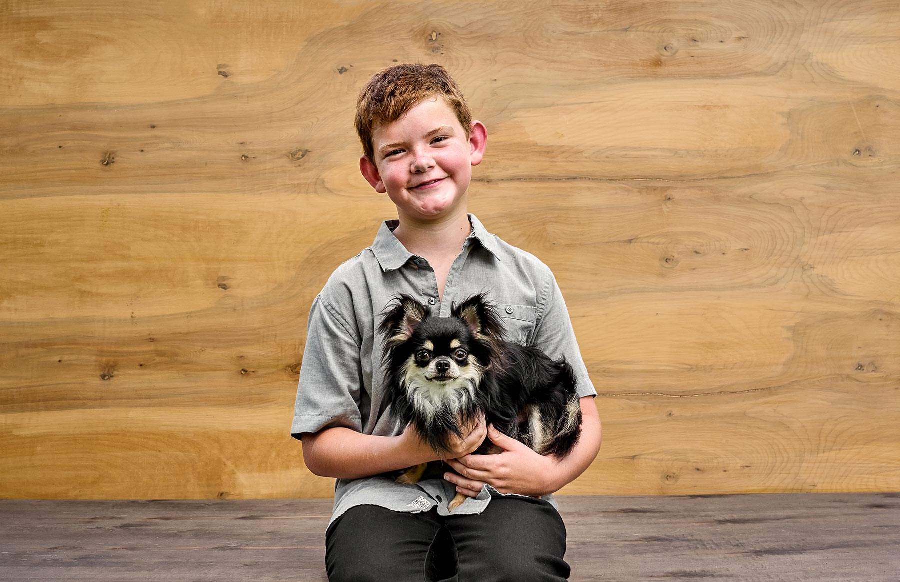 A-young-boy-holding-a-fluffy-black-and-white-dog-against-a-wooden-background