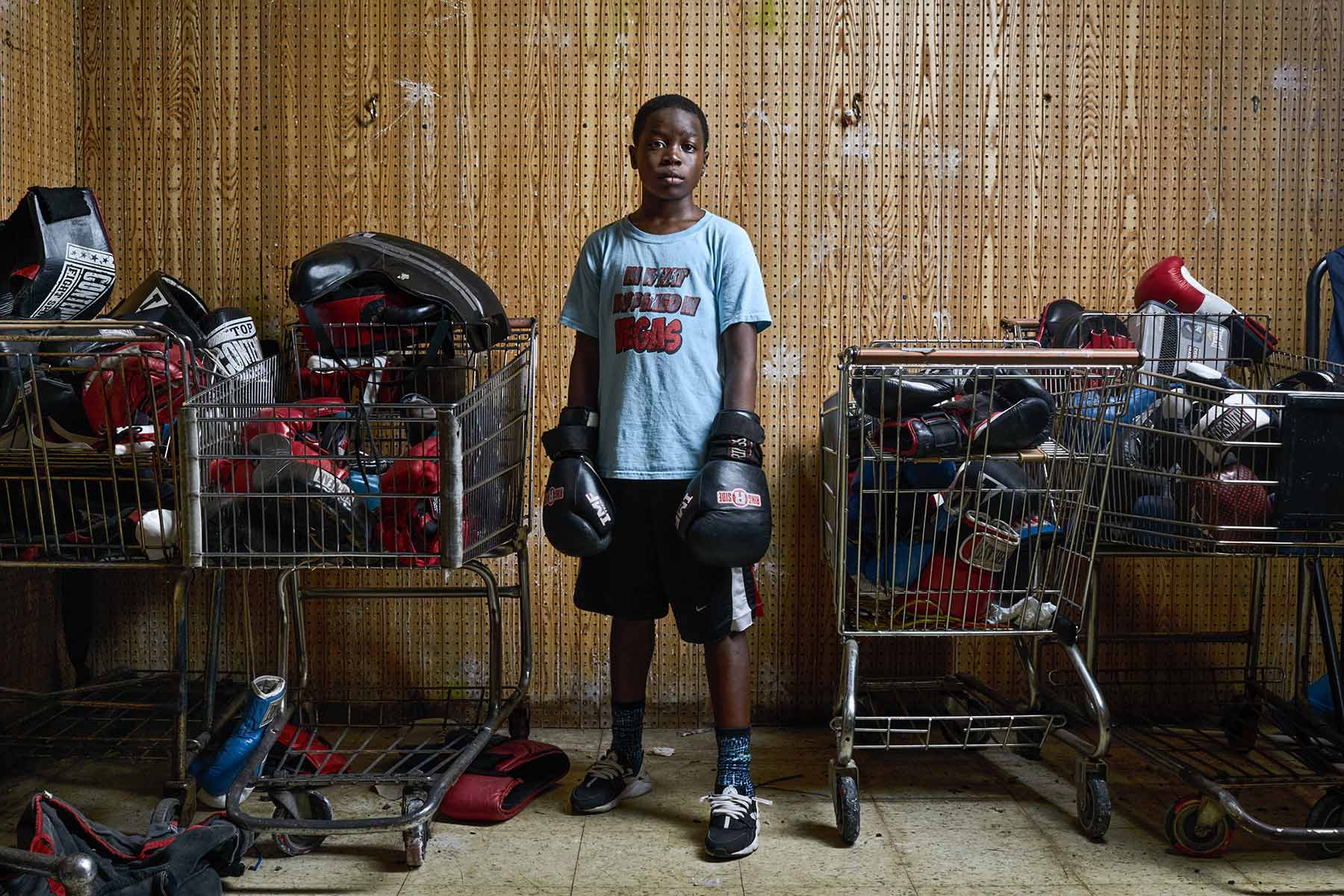 A-portrait-of-a-young-boy-wearing-boxing-gloves-standing-between-four-grocery-carts-full-of-sports-gear