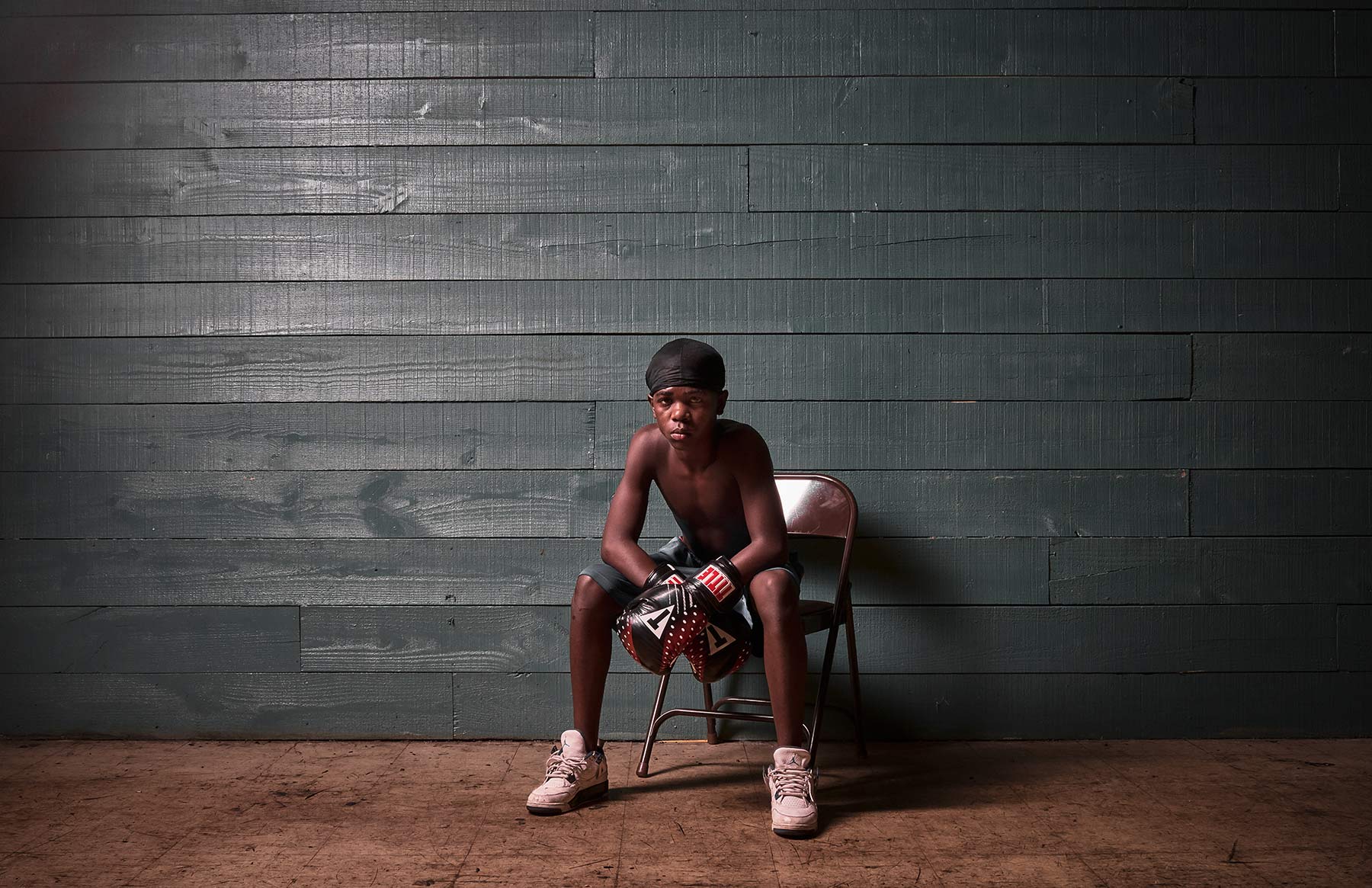 A-portrait-of-a-young-boy-wearing-boxing-gloves-sitting-in-a-chair-in-the-middle-of-an-empty-room-with-panneled-walls